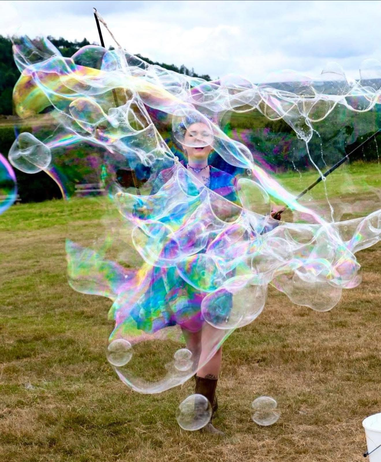 A performer laughing inside a swirling curtain of giant bubbles in an open field