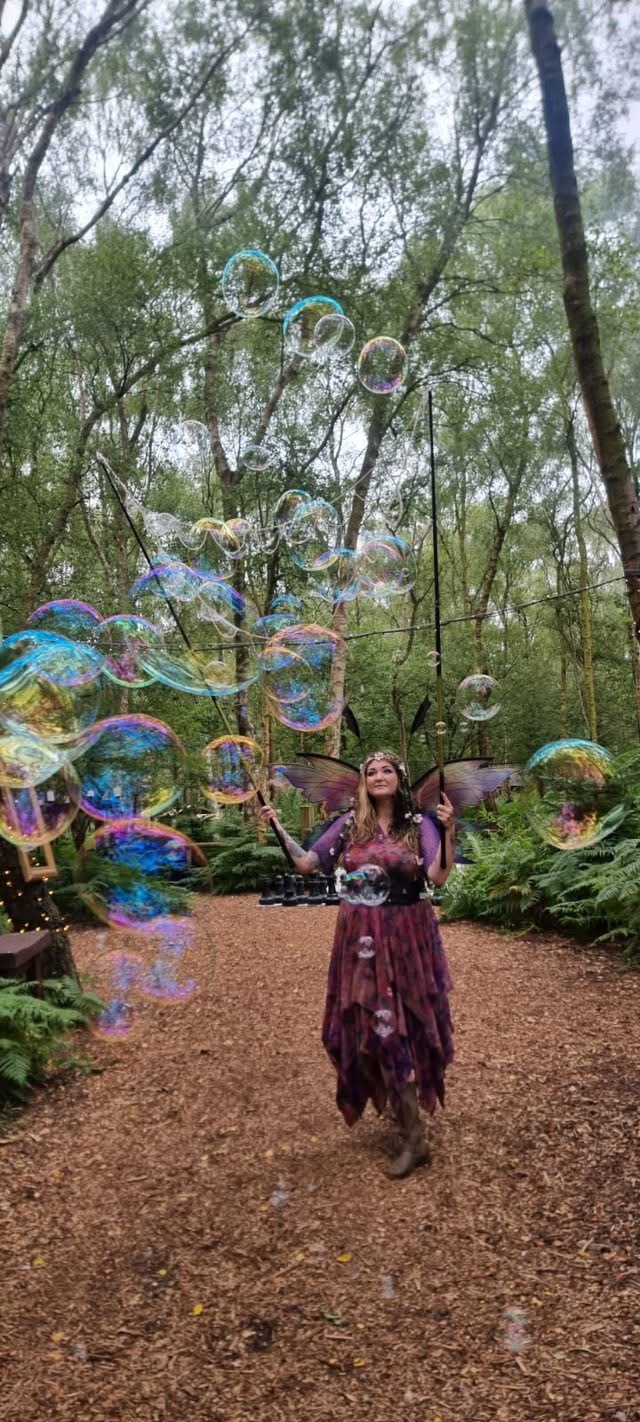 A fairy character on a woodland path beneath a drifting cloud of giant bubbles