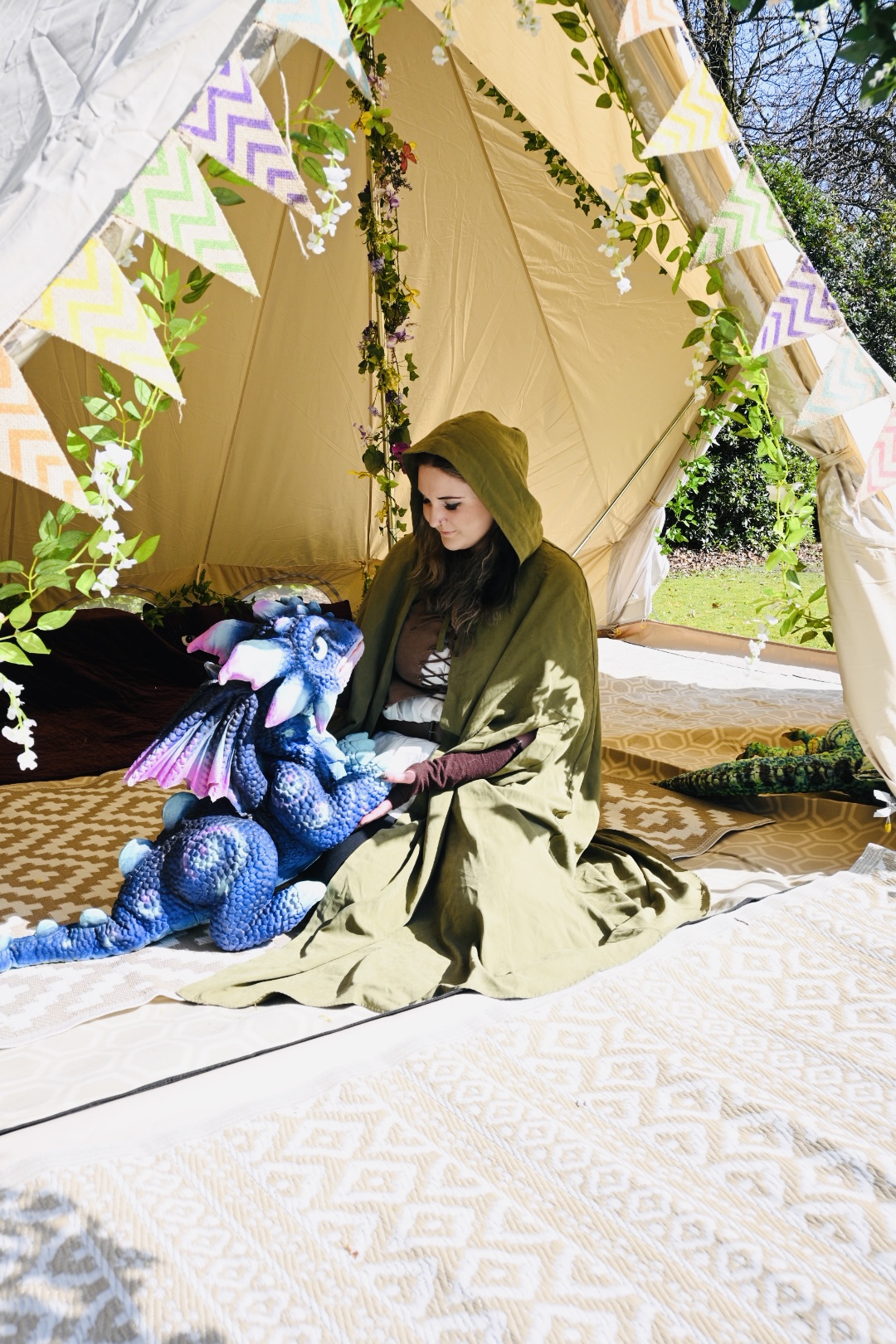 A cloaked storyteller sitting with a blue baby dragon inside a bell tent dressed with bunting and blossom