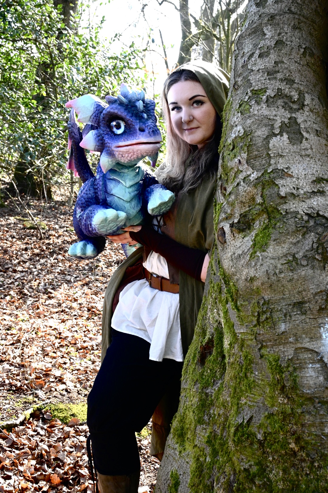 A hooded ranger cradling a blue baby dragon beside a moss-covered tree