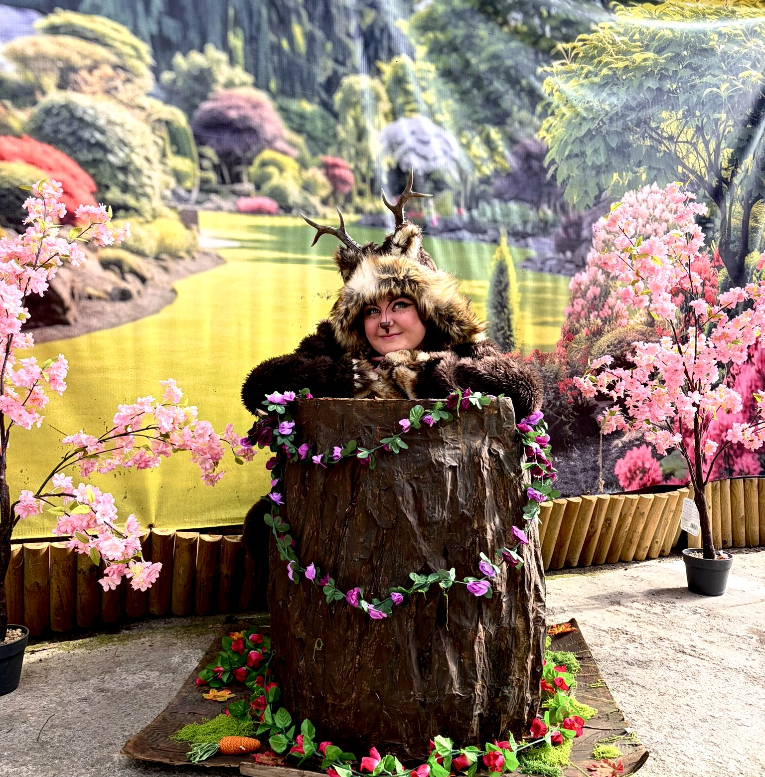 An antlered woodland character in a hollow tree-stump photo booth surrounded by blossom